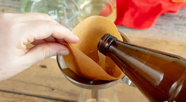 Flüssigkeit wird aus einer braunen Flasche durch einen Kaffeefilter in ein Glas gegossen. So gelingt essig selber machen.