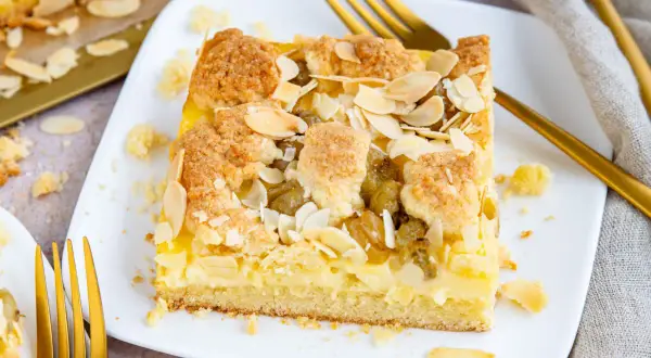 Ein Stück veganer Stachelbeerkuchen mit Streuseln und Mandelblättchen auf einem weißen Teller, daneben goldene Gabeln.