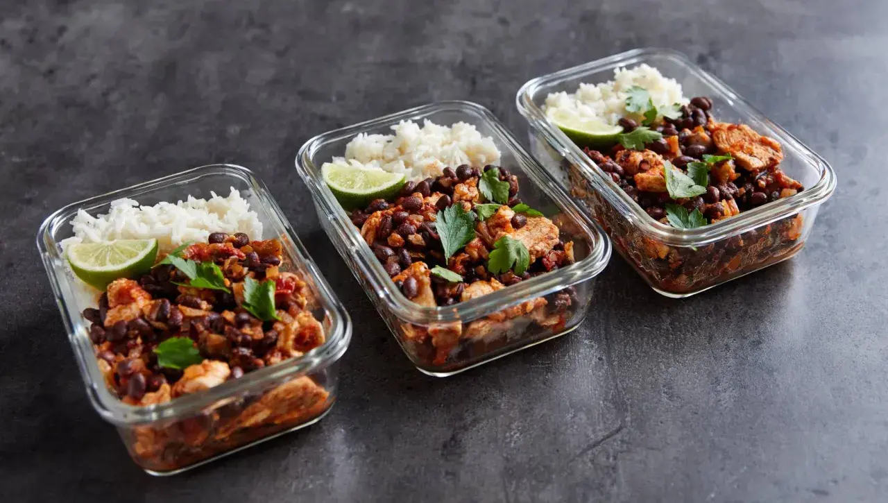Drei Glasbehälter mit meal prep reis, Hähnchen, schwarzen Bohnen und Limettenspalten, garniert mit Petersilie.