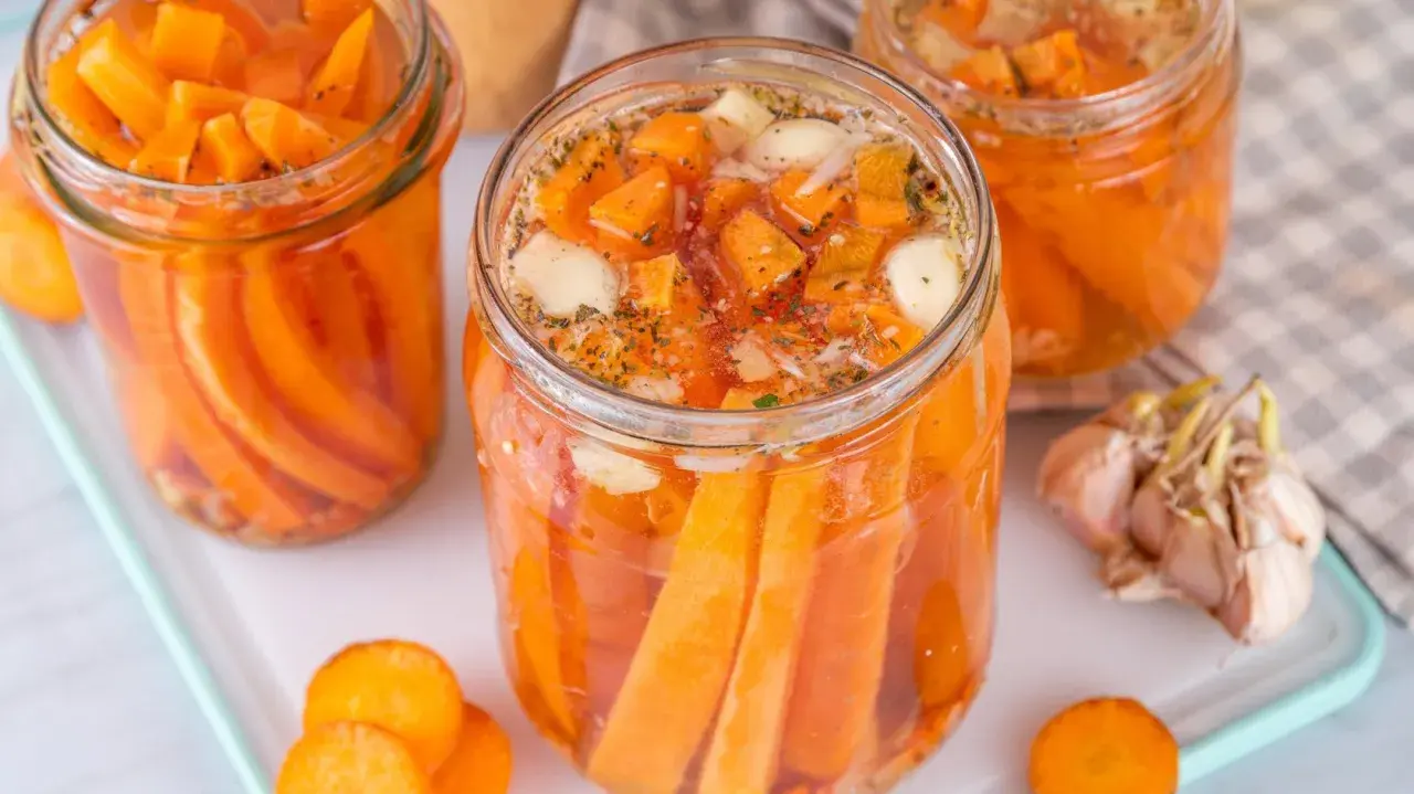 Gläser mit karotten fermentieren, gefüllt mit Karottenstücken, Knoblauch und Kräutern. Daneben liegen frische Karottenscheiben und eine Knoblauchknolle.