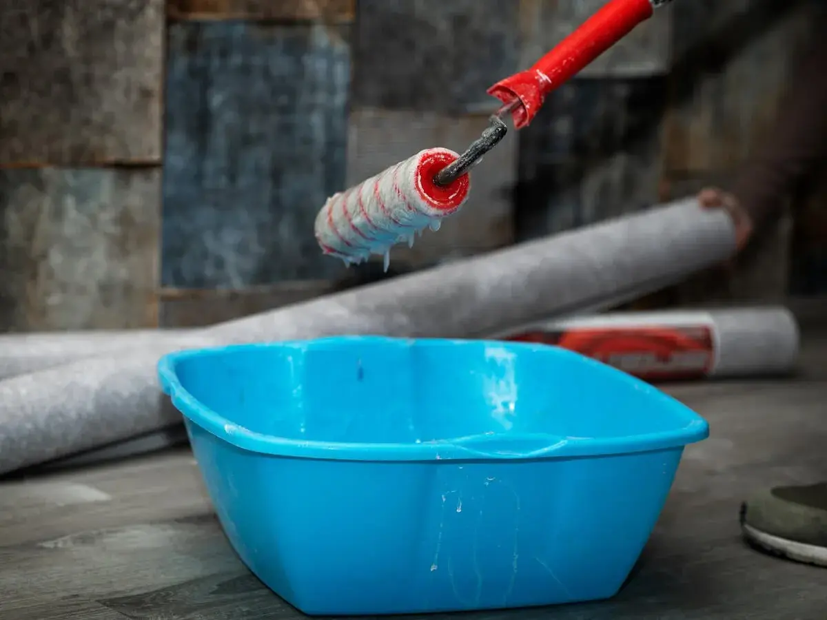Kleister mit idealer kleister konsistenz tropft vom Malerroller in eine blaue Wanne. Tapetenrollen liegen bereit.