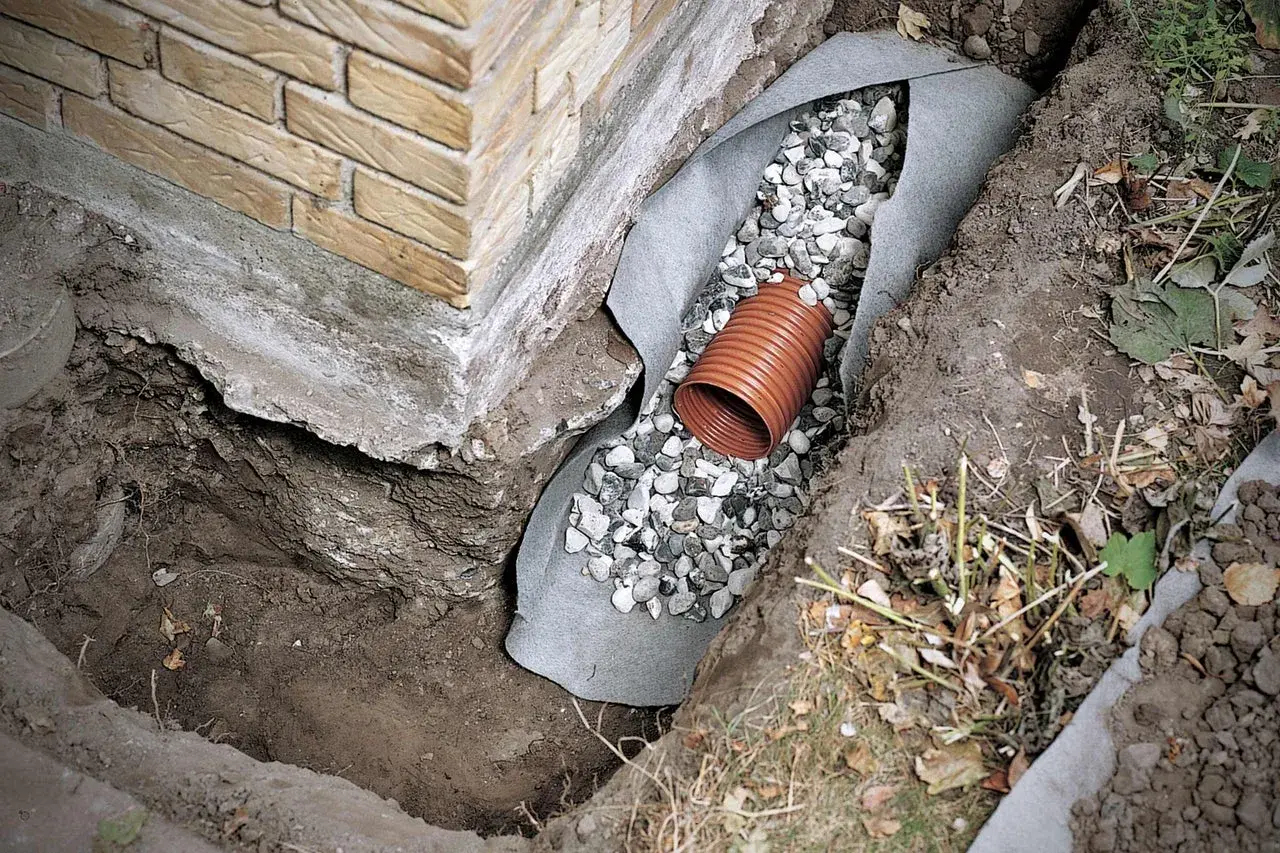 Drainage verlegen: Ein orangefarbenes Rohr liegt in einer Kiesschicht, umgeben von Vlies und Erdreich neben einer Ziegelwand.