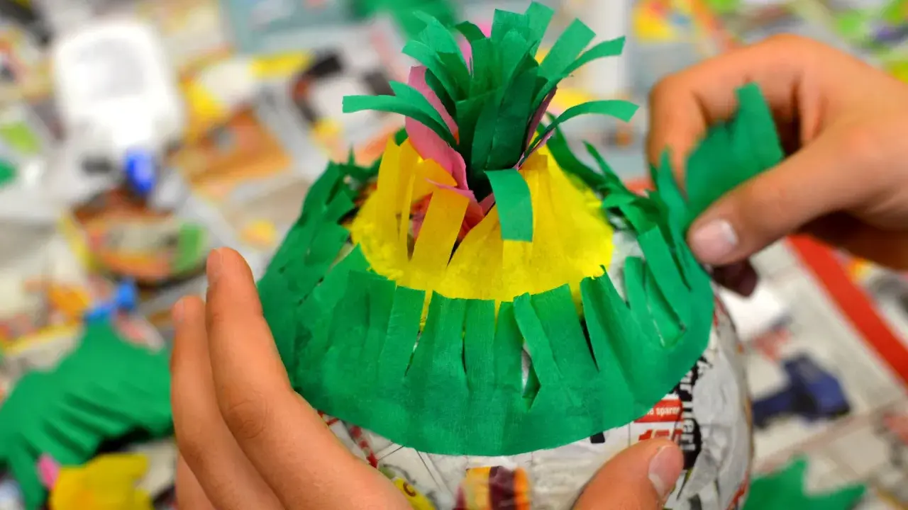 Hände basteln eine bunte Piñata. Mit grünem Krepppapier wird die Form verziert, um eine Piñata selber machen zu können.