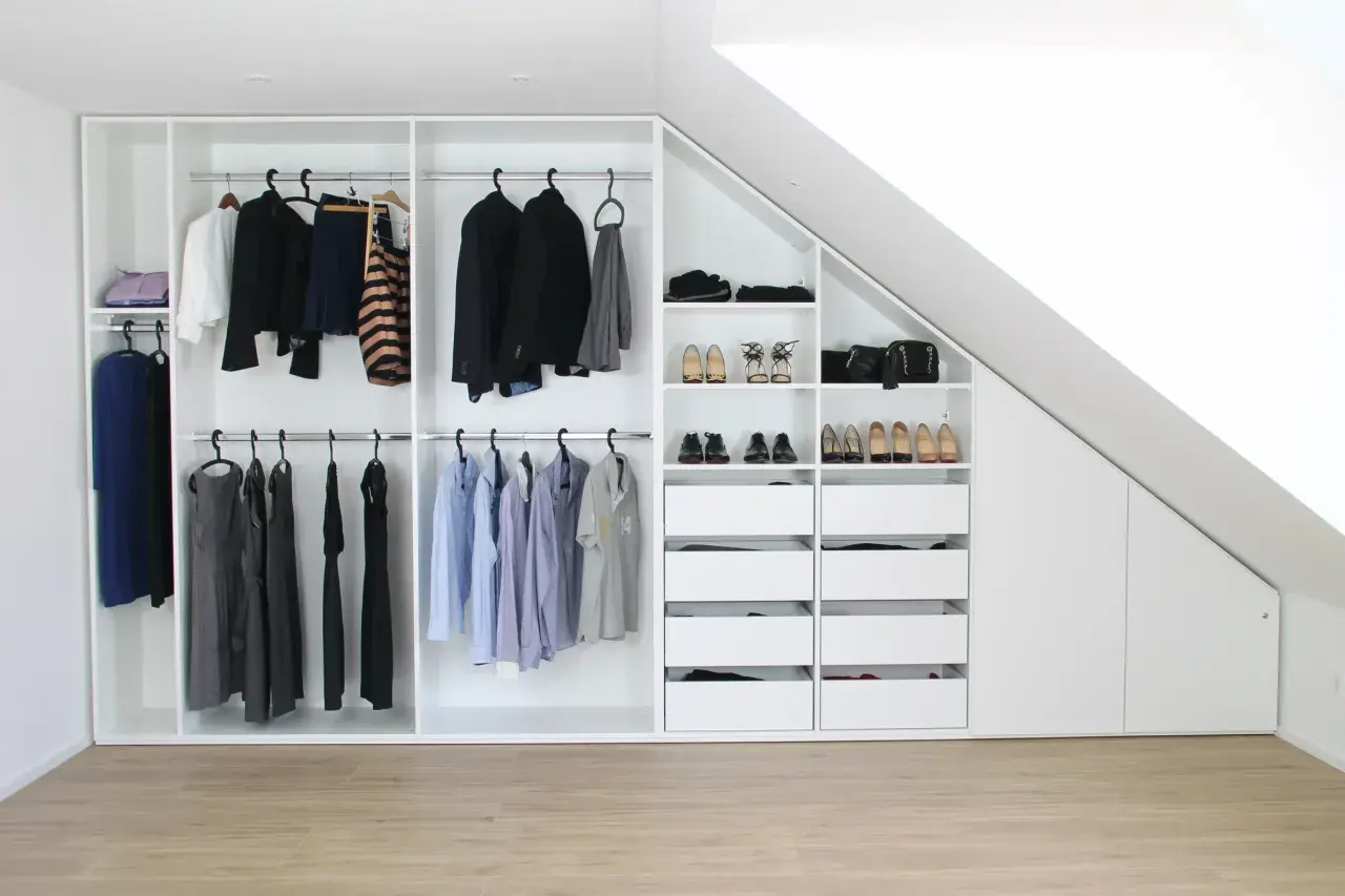 Ein maßgeschneiderter schrank selber bauen: weiße Kleiderschränke mit Kleiderstangen, Schubladen und Regalen, gefüllt mit Kleidung und Schuhen, unter einer Dachschräge.