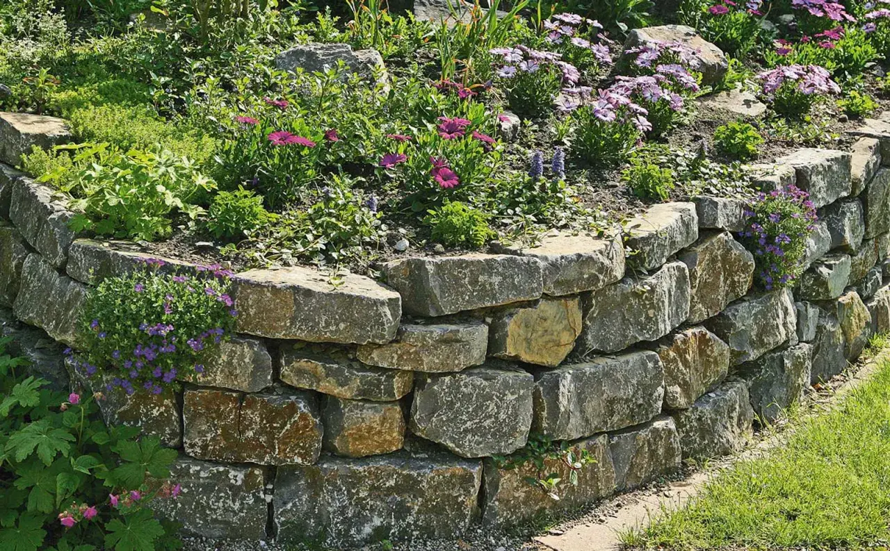 Eine **trockenmauer** sichert den **Hang** mit Natursteinen. Blumen und Grünpflanzen wachsen üppig darauf und daneben.