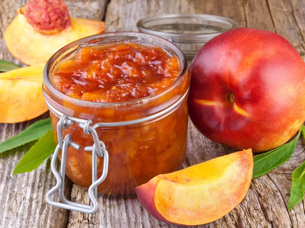 Glas mit leuchtend orangefarbener Nektarinen Marmelade, daneben frische Nektarinen und Fruchtstücke auf rustikalem Holz.