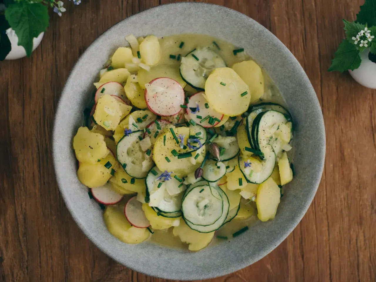Ein Teller mit bayrischem Kartoffelsalat mit Gurke, Radieschen und Schnittlauch, garniert mit kleinen blauen Blüten.