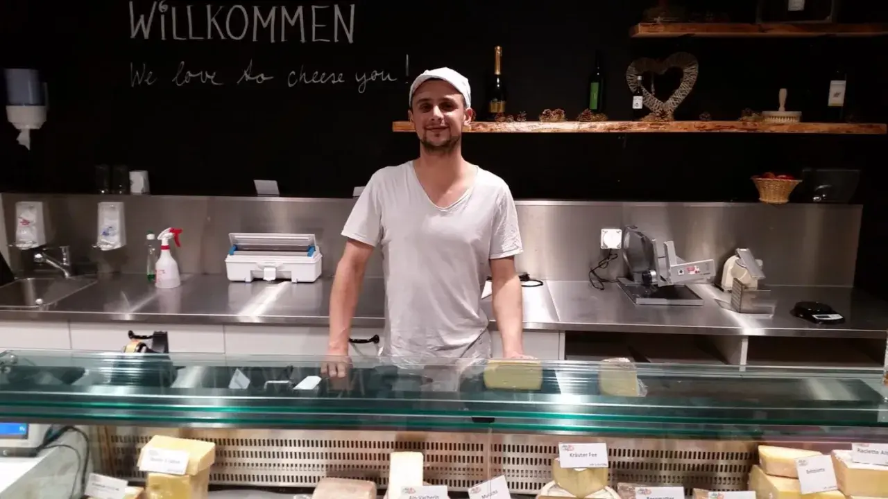 Ein Mann steht hinter einer Käsetheke. Die Tafel über ihm verkündet "Willkommen, we love to cheese you!". Hier gibt es nur das Beste, auch mit tierischem Lab.