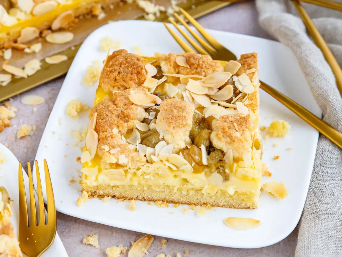 Ein Stück veganer Stachelbeerkuchen mit Streuseln und Mandelblättchen auf einem weißen Teller, daneben goldene Gabeln.