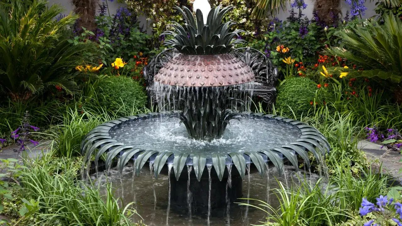 Ein kunstvoller Gartenbrunnen im Ananas-Design, der zum **gartenbrunnen selber bauen** inspiriert. Umgeben von üppiger Vegetation und bunten Blumen.