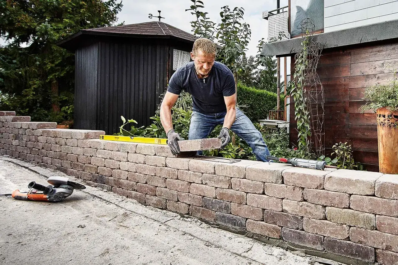 Mann baut eine Gartenmauer aus Ziegelsteinen. Werkzeuge wie Schleifmaschine und Wasserwaage liegen bereit.