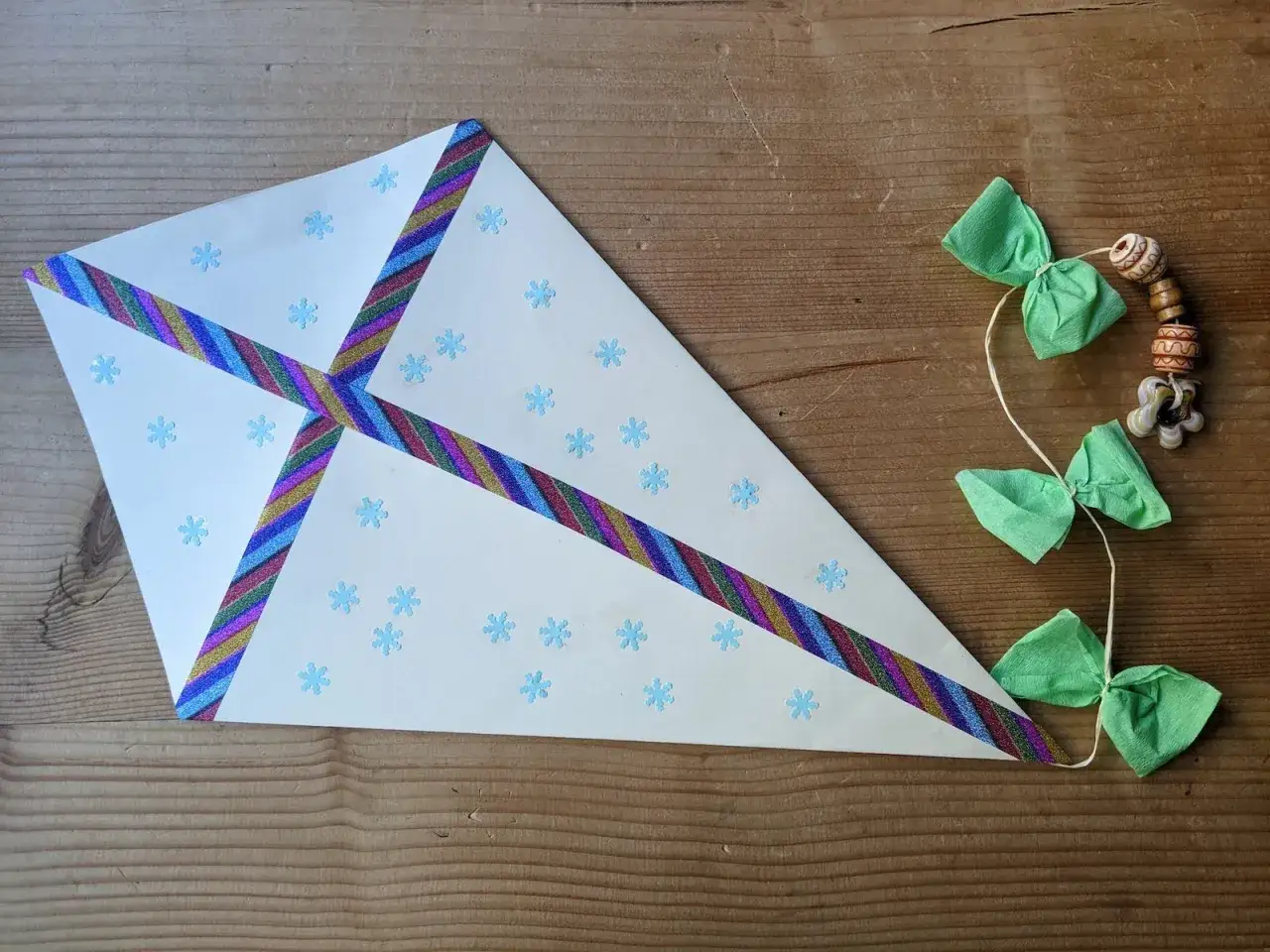 Bastelideen: Ein Drachen aus wei&szlig;em Papier mit Schneeflocken und bunten B&auml;ndern, dazu eine Schnur mit gr&uuml;nen Schleifen und Holzperlen.