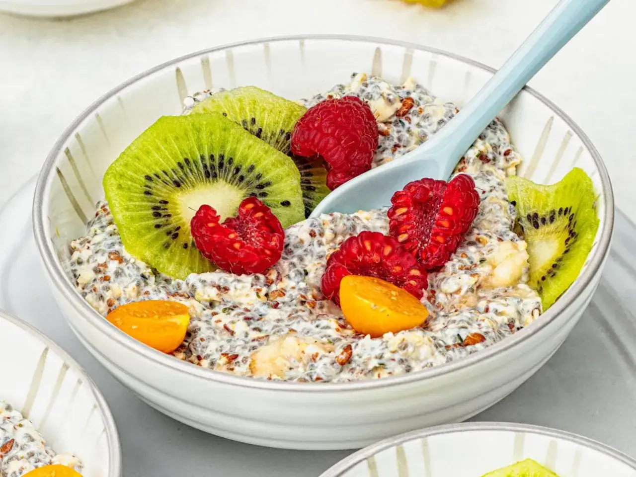 Sch&uuml;ssel mit eingeweichten Haferflocken, garniert mit Kiwi, Himbeeren und Physalis. Ein L&ouml;ffel liegt darin.