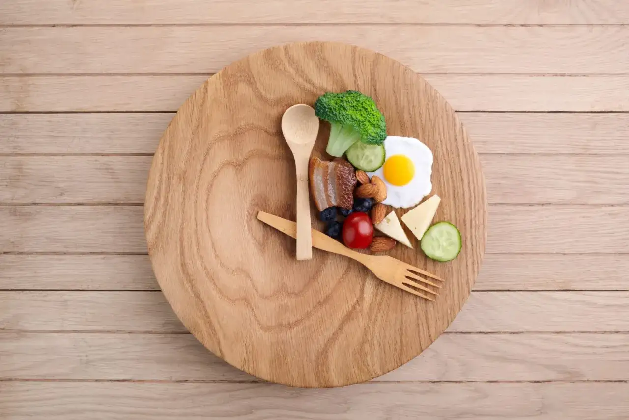 Ein Teller mit einer Mahlzeit, die wie eine Uhr aussieht, symbolisiert 23 Stunden Fasten. Darauf sind Spiegelei, Brokkoli, Gurke, Käse, Mandeln, Blaubeeren, Tomate und Speck.