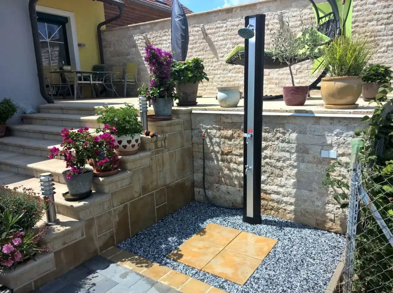 Eine moderne Gartendusche, die man leicht selber bauen kann, steht auf einem Kiesbett mit Steinplatten. Blumen und Pflanzen schm&uuml;cken die Terrasse.
