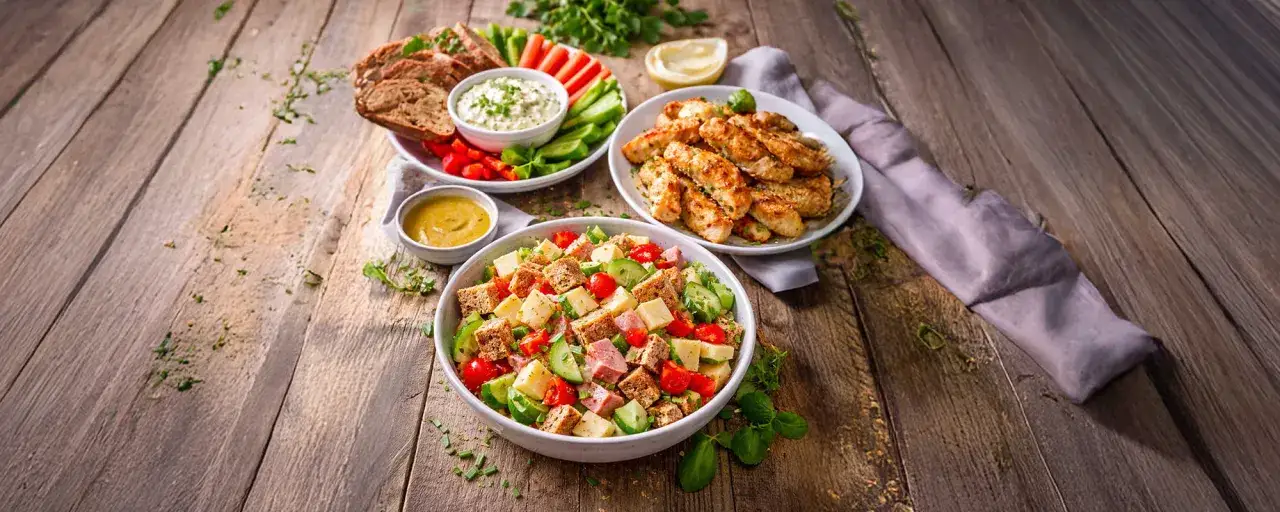 Eine gesunde Brotzeit mit einem bunten Salat, gegrillten Hähnchenstreifen, Brot mit Dip und Gemüsesticks.