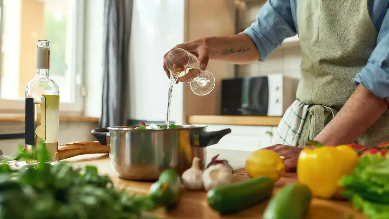 Mann verkocht Wein beim Kochen. Er gie&szlig;t Wei&szlig;wein aus einem Glas in einen Topf mit Gem&uuml;se.