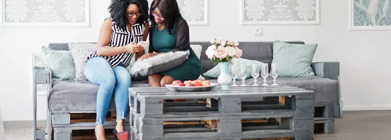 Zwei Frauen lachen &uuml;ber ein Handy, w&auml;hrend sie auf einem selbstgebauten Palettensofa sitzen. Der Couchtisch ist ebenfalls aus Paletten gebaut.