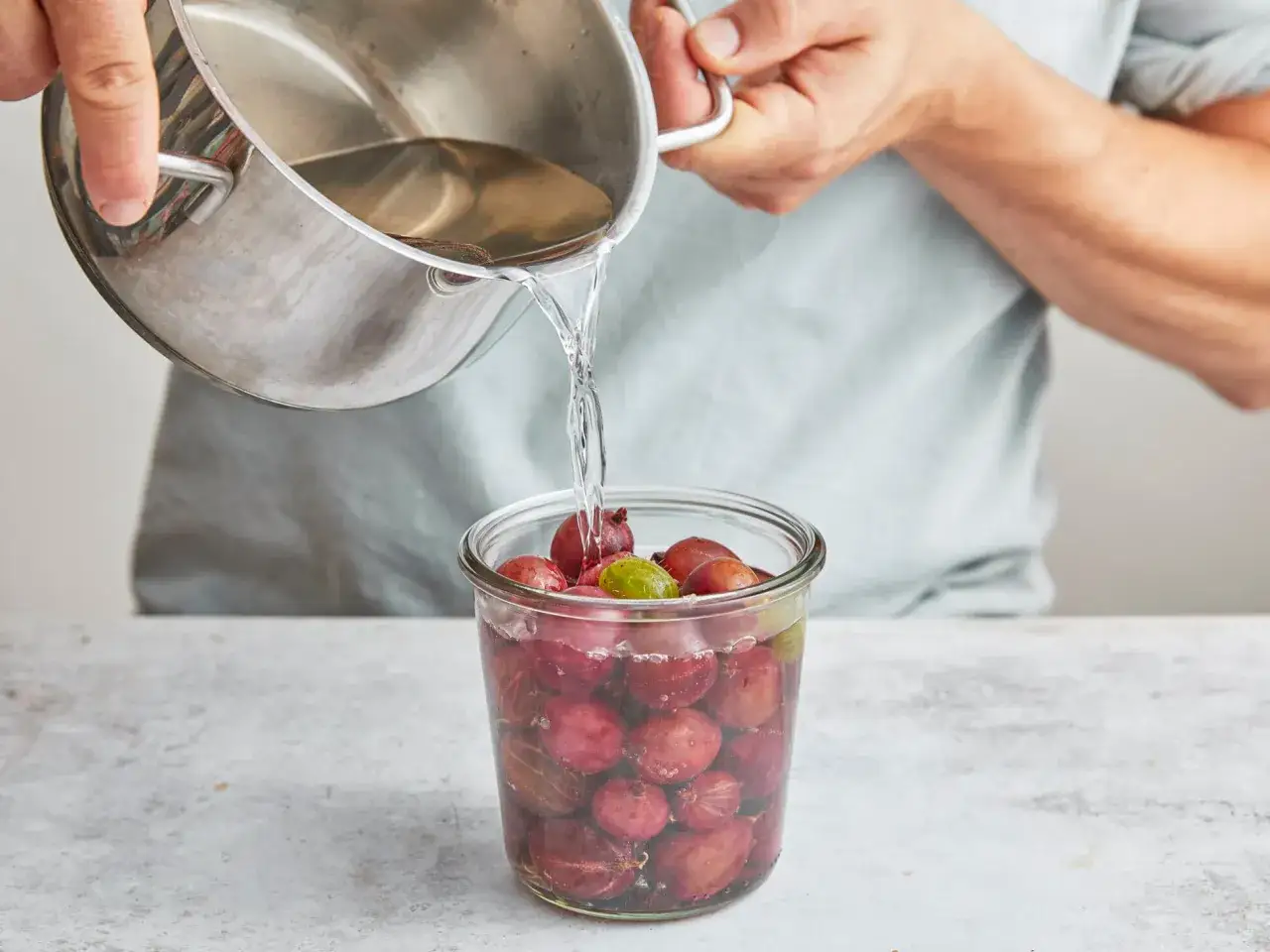 Flüssigkeit wird aus einem Topf in ein Glas voller roter und grüner Stachelbeeren gegossen. So werden die Früchte für die Lagerung vorbereitet.