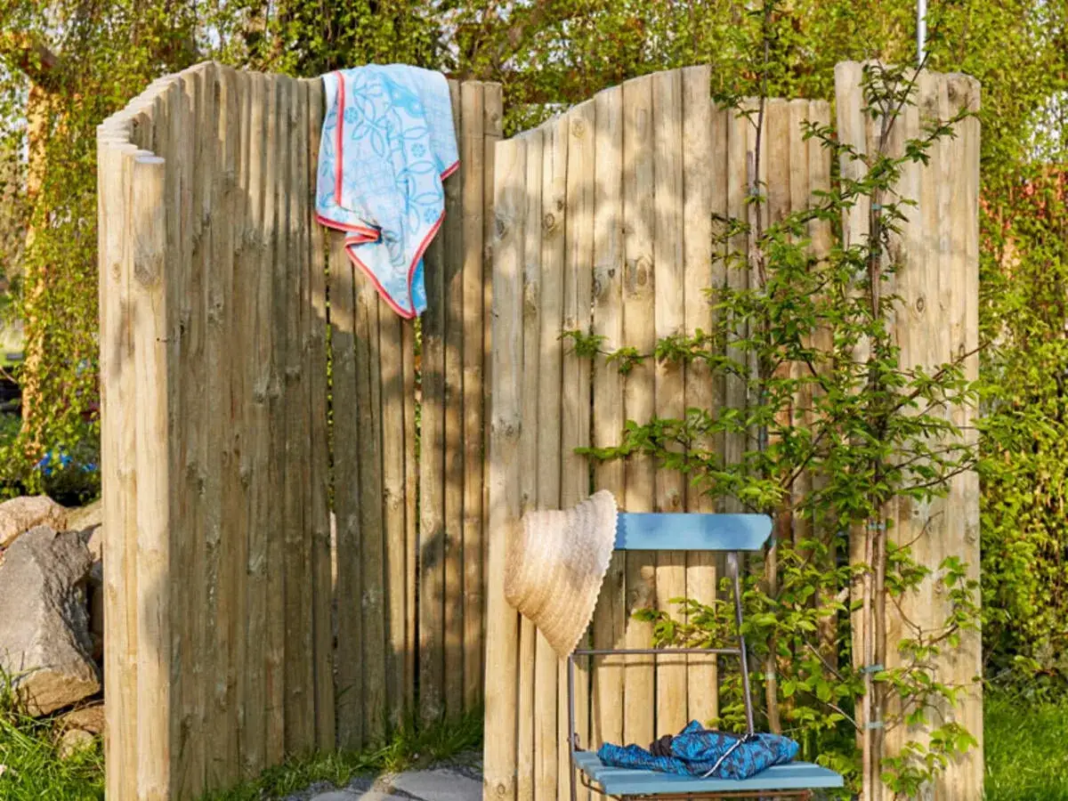 Eine rustikale Holzwand, perfekt f&uuml;r eine Gartendusche selber bauen. Ein Handtuch h&auml;ngt, ein Strohhut und eine blaue Decke liegen auf einem Stuhl.