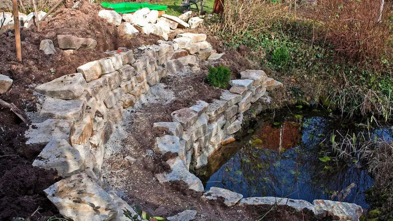Eine neu errichtete trockenmauer hangbefestigung aus Natursteinen sichert den Uferbereich eines kleinen Teichs.