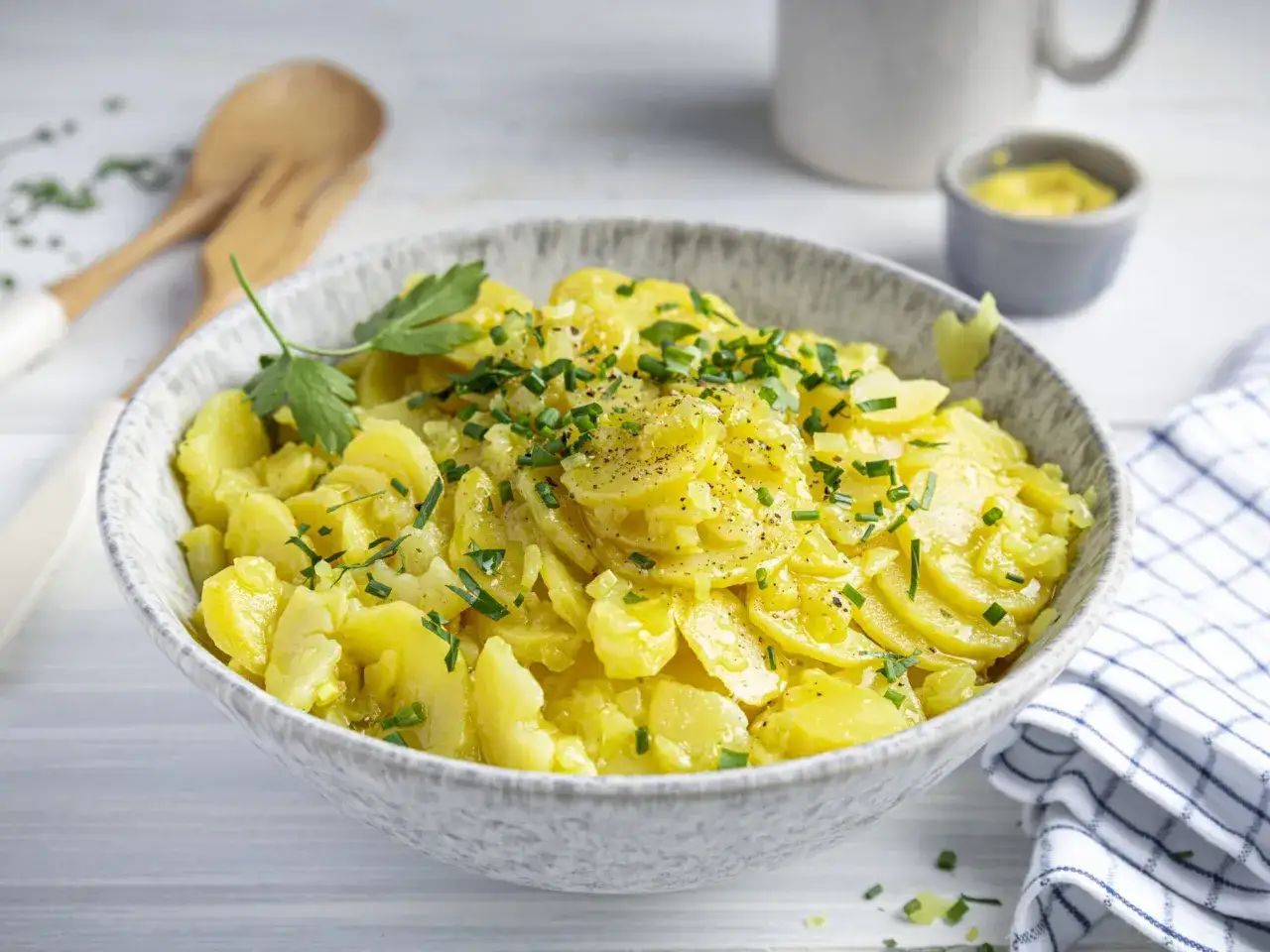 Schüssel mit bayrischem Kartoffelsalat mit Gurke, garniert mit Schnittlauch und Petersilie. Daneben ein kleines Schälchen mit Senf.