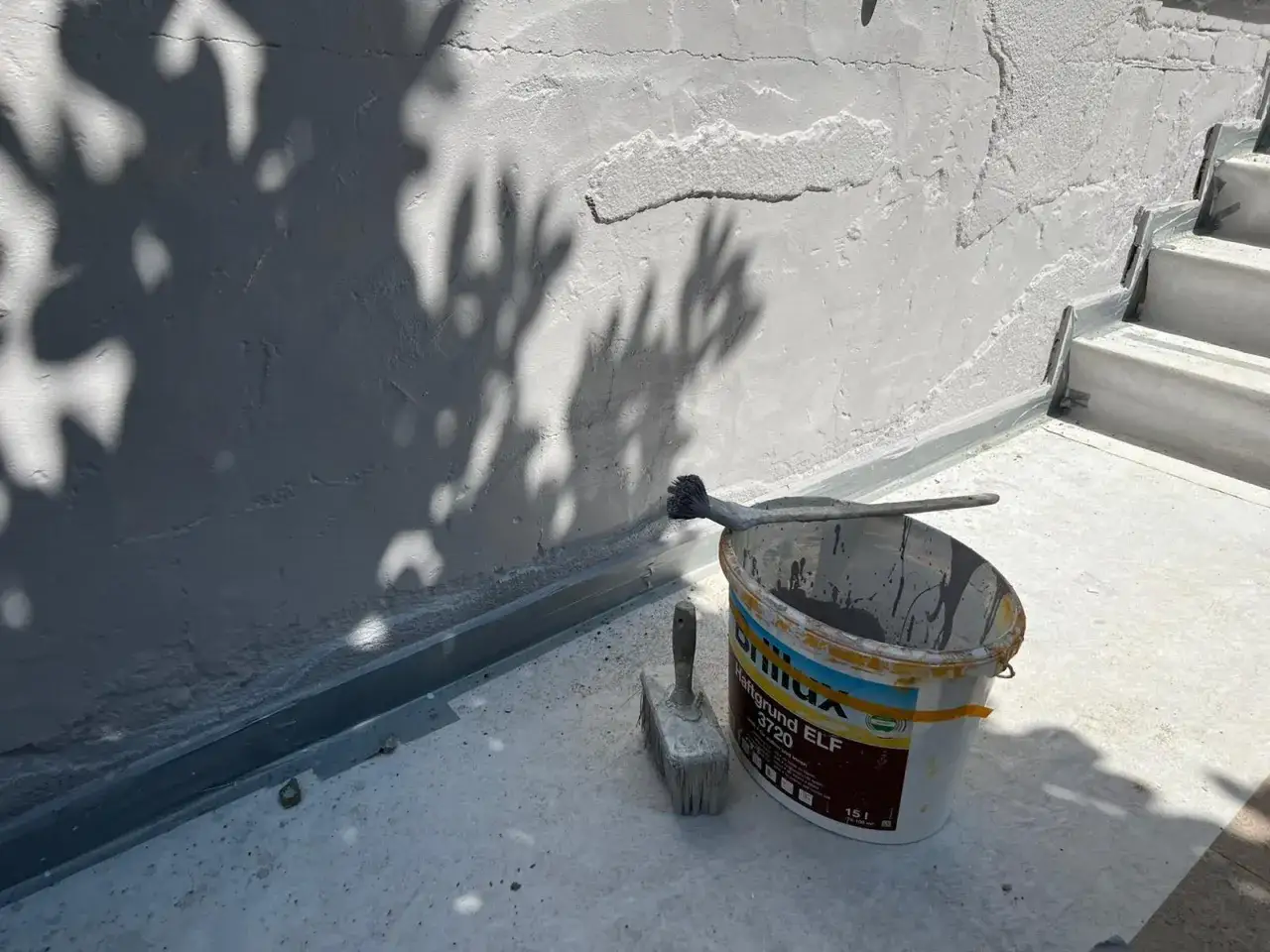 Werkzeug und Farbeimer bereit zum Sockel verputzen. Schatten von Blättern tanzen auf der Wand.