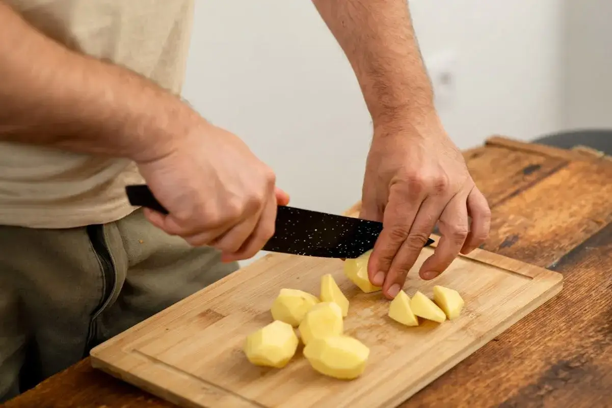 H&auml;nde schneiden rohe Kartoffeln auf einem Holzbrett. Manche Leute essen Kartoffeln roh, aber das ist nicht &uuml;blich.
