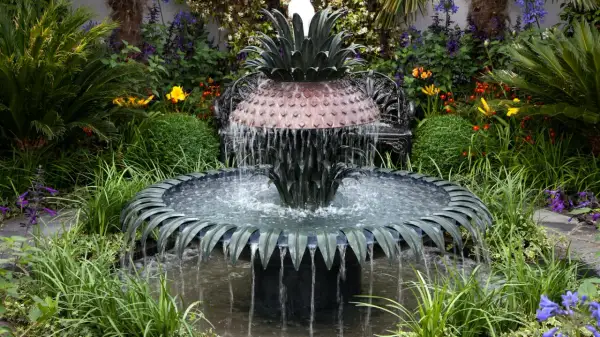 Ein kunstvoller Gartenbrunnen im Ananas-Design, der zum **gartenbrunnen selber bauen** inspiriert. Umgeben von üppiger Vegetation und bunten Blumen.