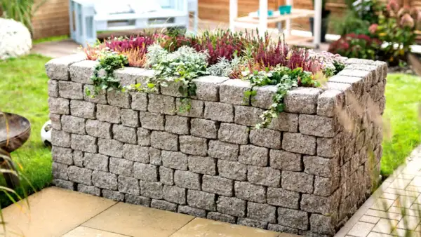 Ein bepflanztes Hochbeet mit stabilen hochbeet mauern aus Betonsteinen, umgeben von grünem Rasen und einer Terrasse.