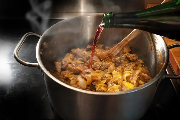 Rotwein wird beim Kochen in einen Topf mit Fleisch und Zwiebeln gegossen. Dampf steigt auf, während das Gericht köchelt.