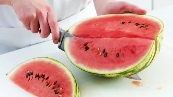 So schneidest du eine Wassermelone richtig: Ein Messer teilt die saftige Frucht in zwei Hälften, eine davon liegt bereits als Stück daneben.