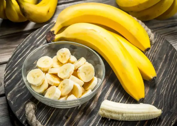 Bananen auf einem Holzteller, eine Schüssel mit Bananenscheiben und eine geschälte Banane. Interessiert, wie viel Zucker hat eine Banane?