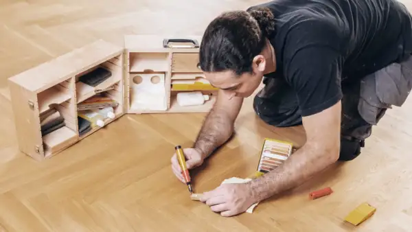 Mann repariert Parkett mit einem Reparaturstift und einer Farbkarte. Werkzeugkasten mit Zubehör daneben.