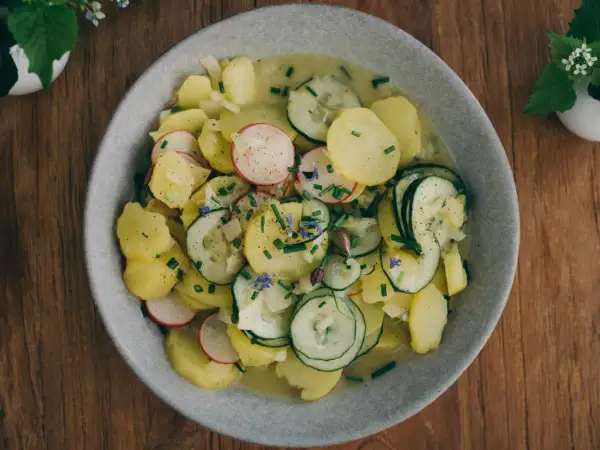 Ein Teller mit bayrischem Kartoffelsalat mit Gurke, Radieschen und Schnittlauch, garniert mit kleinen blauen Blüten.
