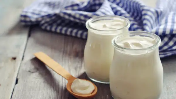 Zwei Gläser cremigen griechischen Joghurts, reich an Protein, stehen auf einem Holztisch. Ein Holzlöffel mit einer Portion Joghurt liegt daneben.